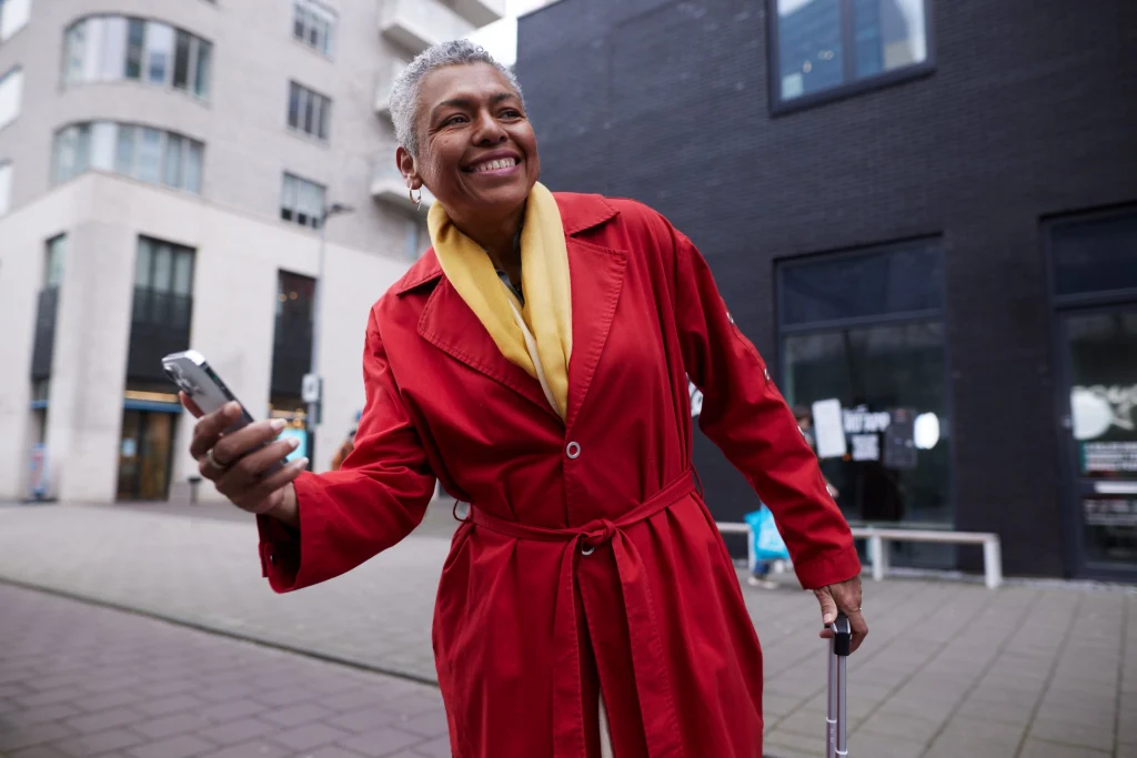 Lachende vrouw in rode jas met telefoon en koffer in de stad, campagnebeeld voor TomTom